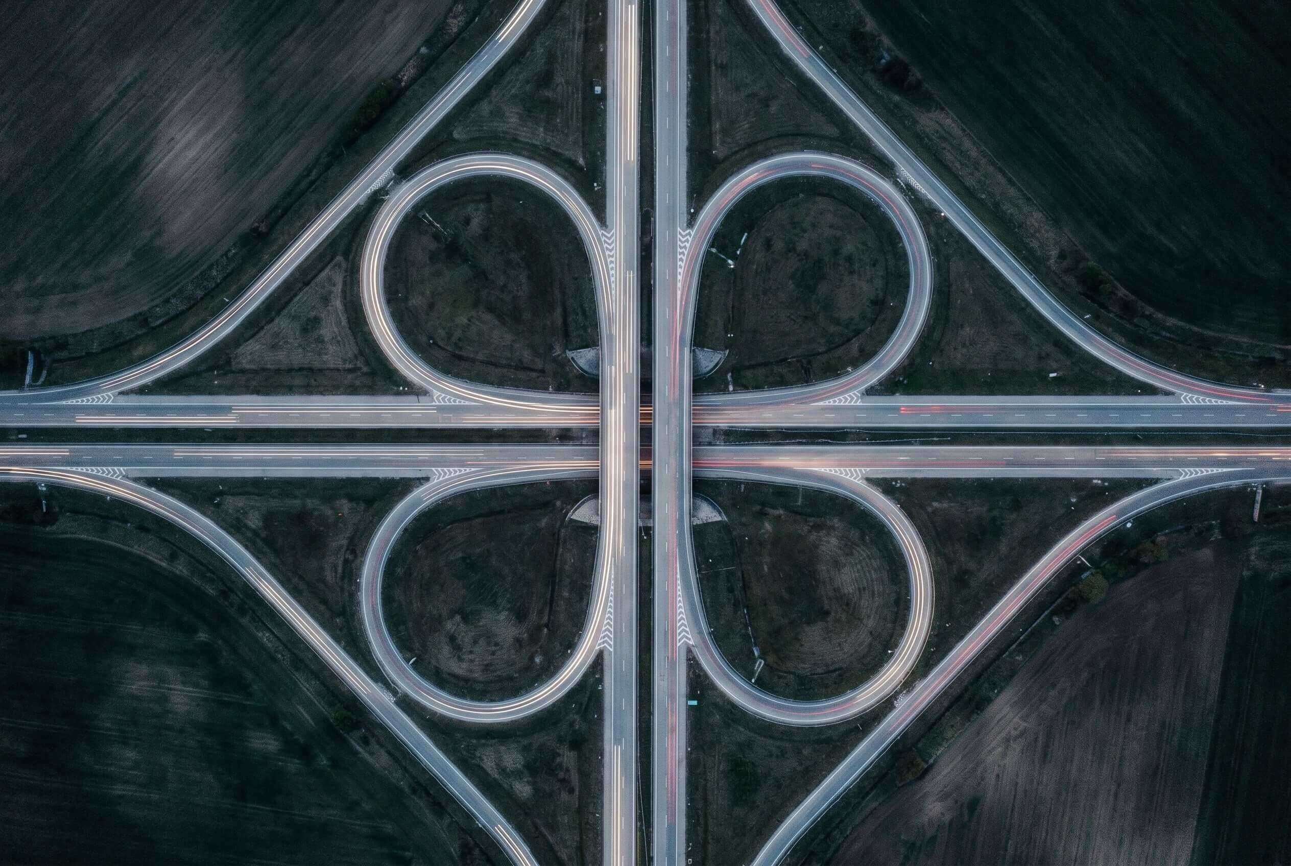 A cloverleaf motorway interchange from above at blue hour, light trails tracing the loops