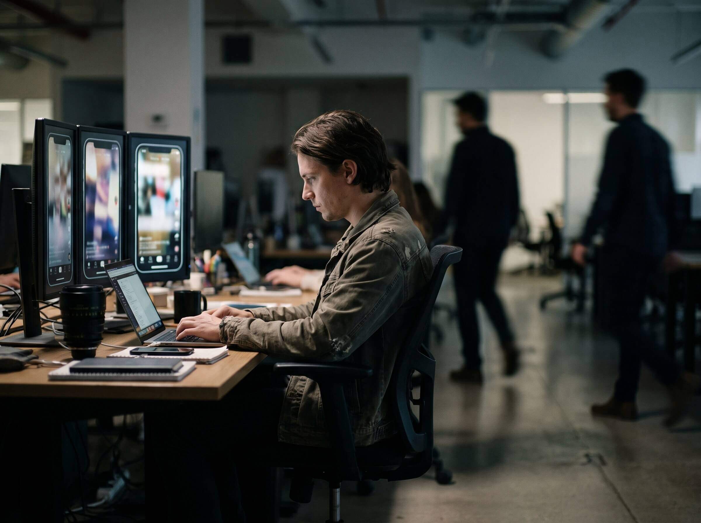 A social media editor at a desk with vertical screens showing short video posts, colleagues walking behind him